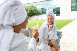 Young girlfriends relaxing in white bathrobes and drinking healthy spa cocktails outside at a resort hotel while talking. Furniture, with hotel landscaping in the background.