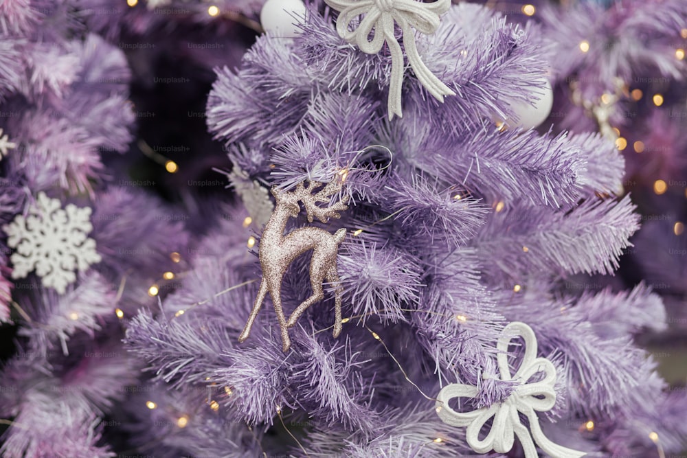 Brinquedo de rena de purpurina elegante na árvore de Natal roxa moderna com ornamentos e luzes na frente da loja ou na fachada do edifício. Decoração de rua festiva de Natal para férias de inverno. Feliz Natal