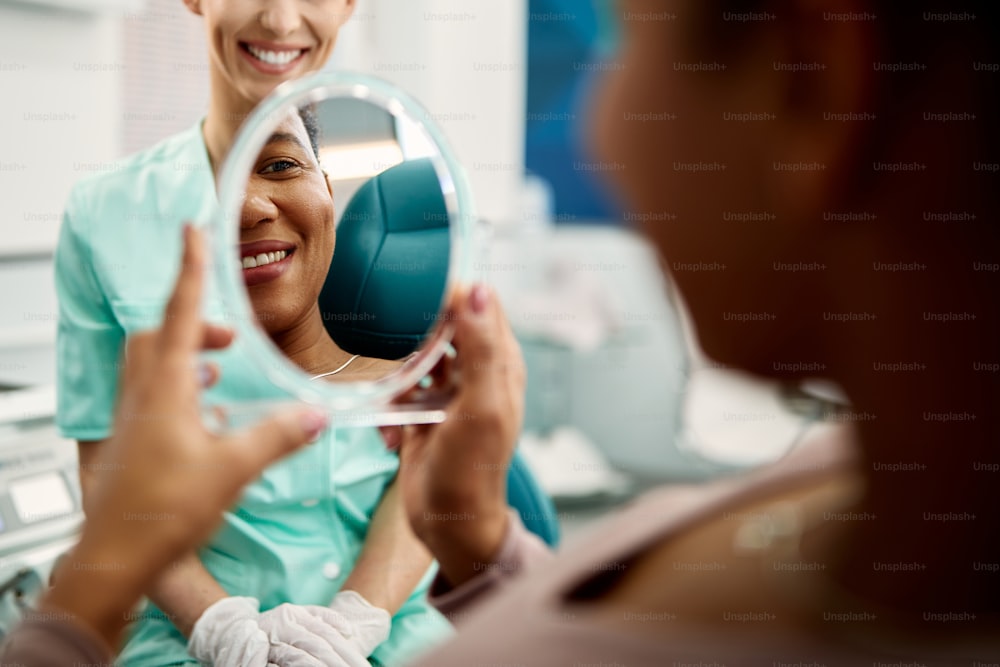 Close-up of satisfied African American woman using mirror and looking at her teeth at dentist's office.