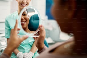 Primer plano de una mujer afroamericana satisfecha usando un espejo y mirando sus dientes en el consultorio del dentista.