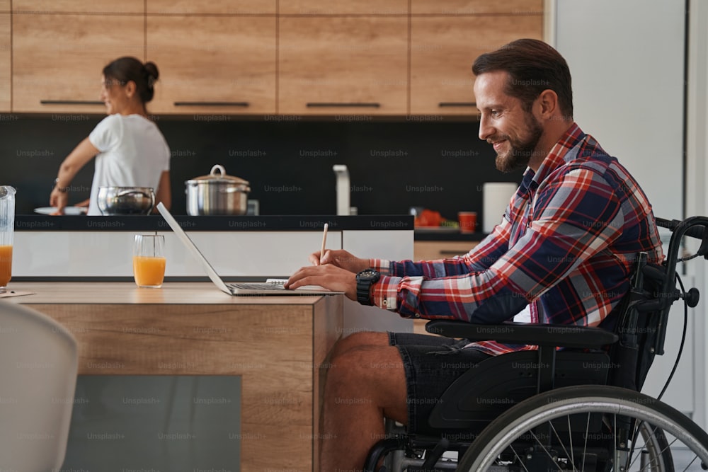 Uomo bello felice che usa una sedia a rotelle mentre lavora online dall'ufficio di casa