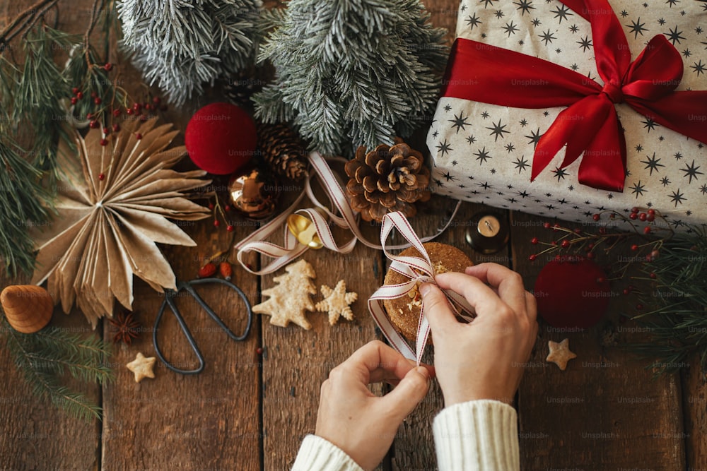 Feliz Natal! Mãos amarrando fita em biscoitos de aveia de Natal no fundo de decorações festivas elegantes na mesa de madeira rústica. Imagem atmosférica de Natal com biscoitos saudáveis e decoração de Natal
