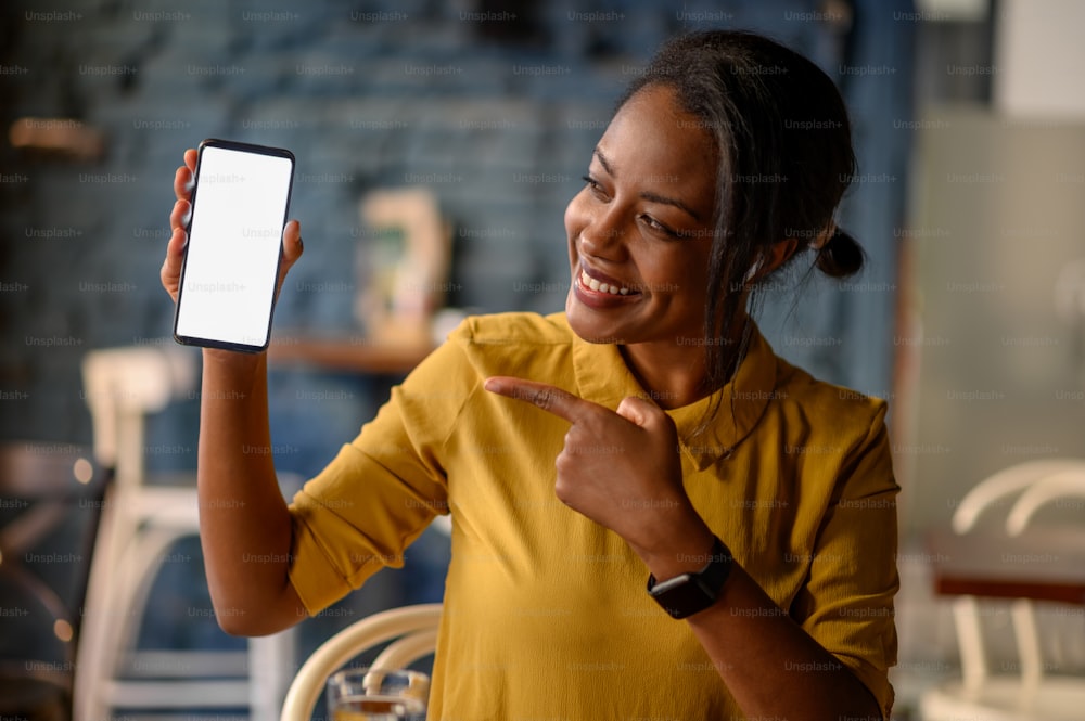 Mulher afro-americana jovem bonita que mostra um smartphone enquanto está sentado em um café