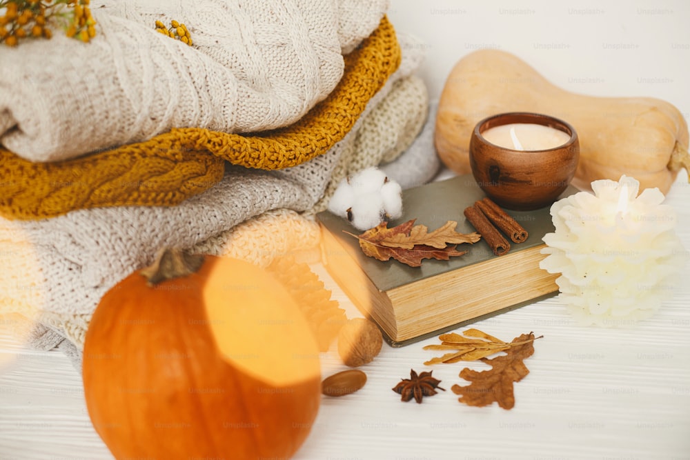 Hello autumn, cozy slow living. Pumpkin, cozy sweaters, autumn leaves, burning candle and vintage book in golden lights bokeh on white wooden background in room.  Happy Thanksgiving