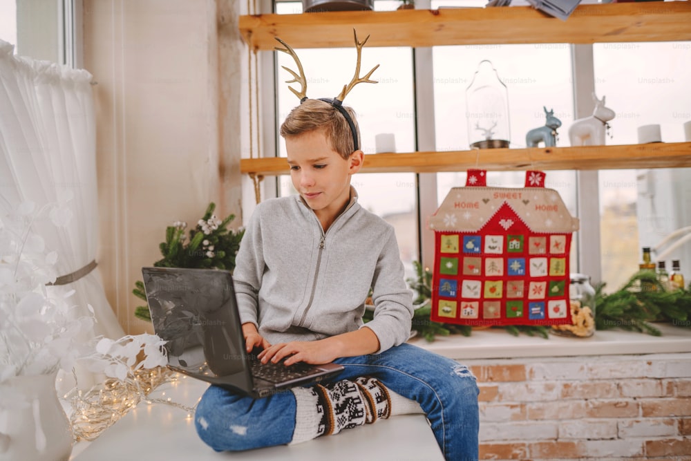 鹿の角のカチューシャをつけた男の子が、屋内のクリスマスの手作りのアドベントカレンダーの近くに座っているラップトップを使用しています。