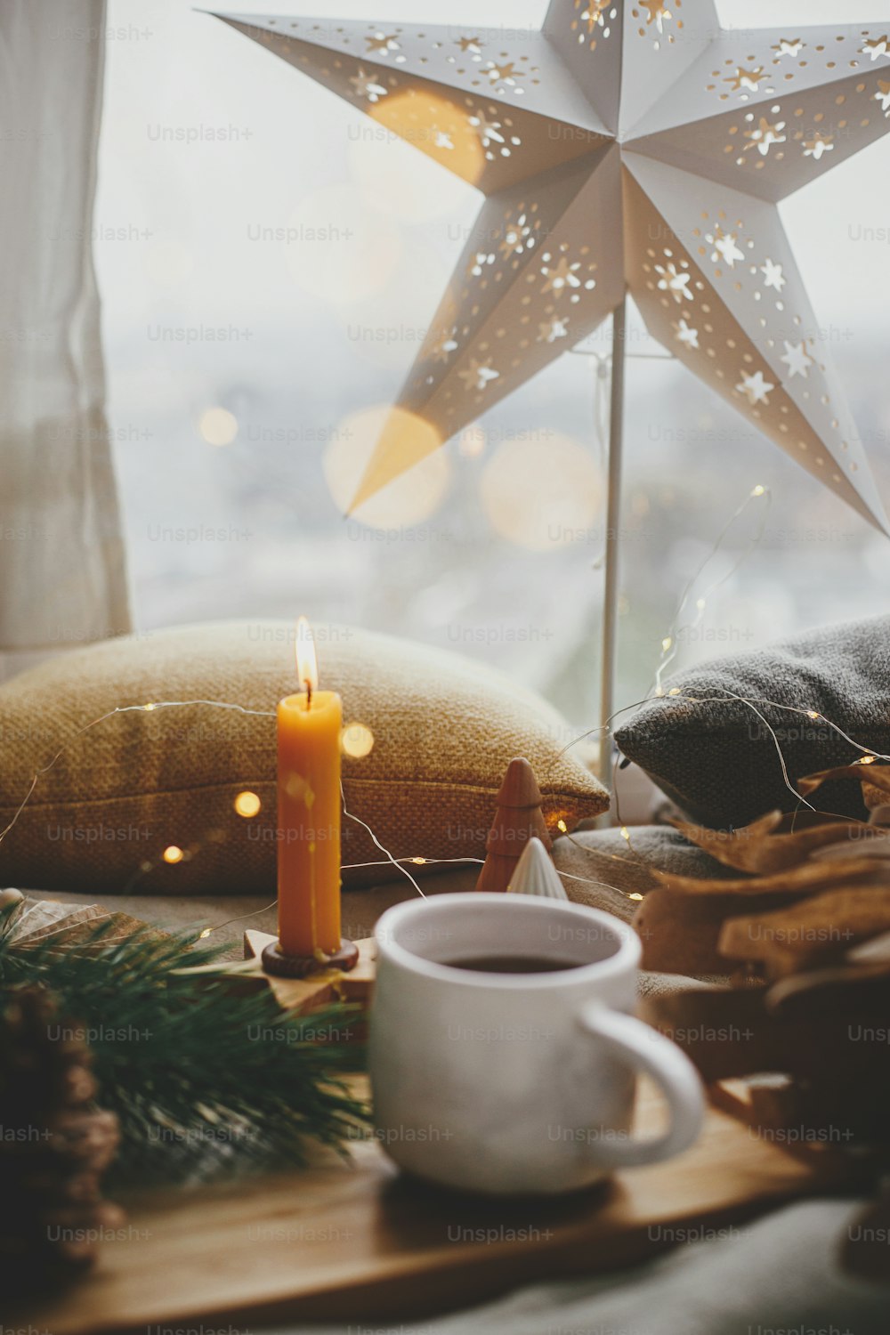 Beleuchteter Weihnachtsstern auf dem Hintergrund einer warmen Tasse Tee, Weihnachtsbeleuchtung, Pinien, Kerze, Kissen am Fenster. Skandinavische Winterhygge. Gemütliches Zuhause. Atmosphärischer magischer Moment