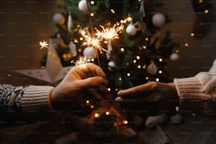 Pareja celebrando con luces de bengala de fuegos artificiales en el fondo del árbol de Navidad y la estrella brillante. ¡Feliz Año Nuevo! Manos sosteniendo bengalas encendidas en una sala festiva escandinava. Momento atmosférico