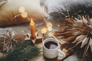 Warm cup of tea on soft bed with christmas stars, golden lights, pine trees, candle and pillows in scandinavian room. Cozy home. Atmospheric magic moment. Hygge winter holidays