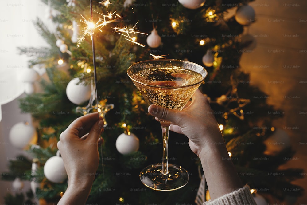 Mani che tengono scintillante ardente e bicchiere di champagne sullo sfondo delle luci dell'albero di Natale. Felice Anno Nuovo! Fuochi d'artificio bengala luce e bere nelle mani della donna. Momento atmosferico. Festa di Capodanno