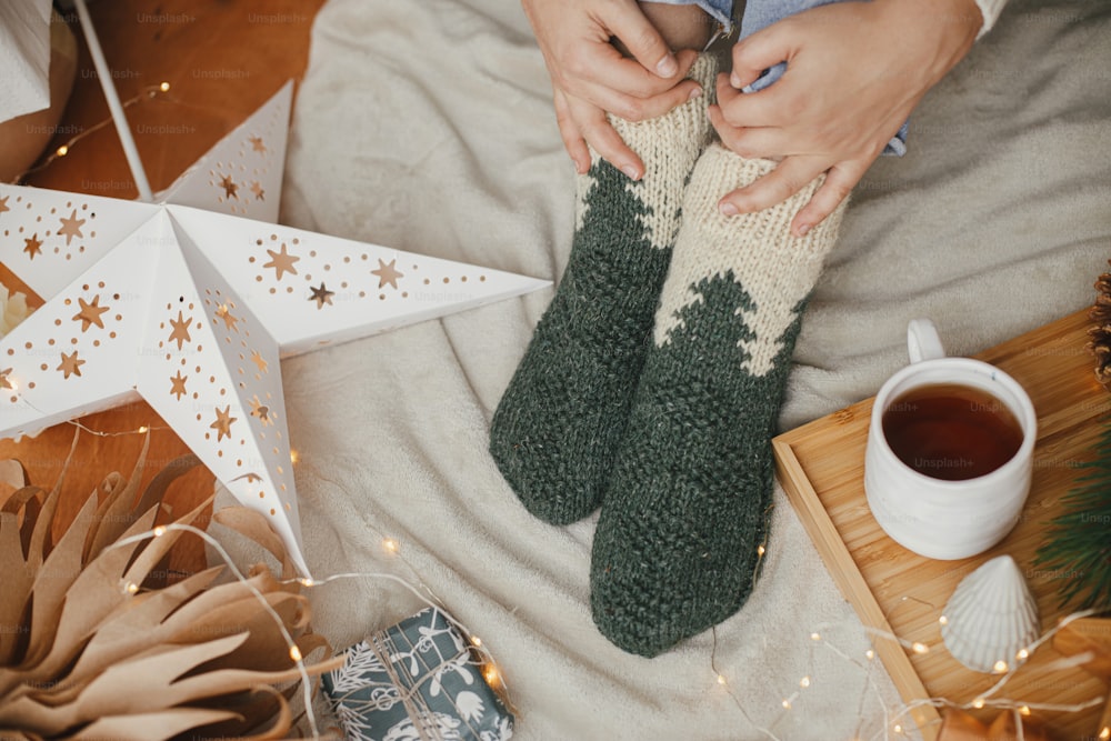 Cozy winter moments at home. Woman feet in woolen socks on soft bed with warm cup of tea, christmas stars, golden lights, trees, candle. Girl relaxing in scandinavian evening room