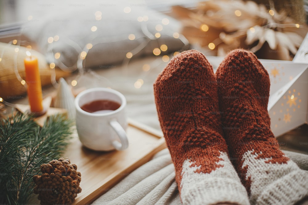 Pies de mujer en calcetines de lana en cama suave con taza de té caliente, estrellas de Navidad, luces doradas, árboles, velas y almohadas. Acogedores momentos invernales en casa. Chica relajándose en la sala de noche escandinava