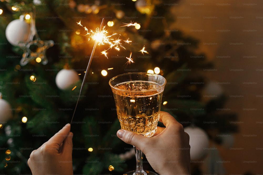 Donna che celebra con luce di fuochi d'artificio e bicchiere di champagne sullo sfondo delle luci dell'albero di Natale. Felice Anno Nuovo! Mani che tengono scintille accese e bevono in una stanza scandinava. Momento atmosferico