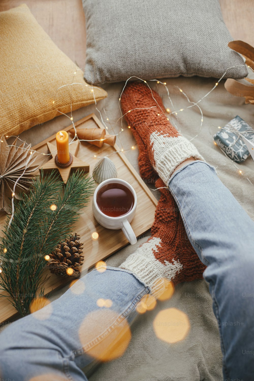 Woman legs in warm socks on soft blanket with cup of tea, christmas stars, golden lights, trees, candle and pillows, top view. Cozy relaxing moments at home. Winter and autumn holidays.