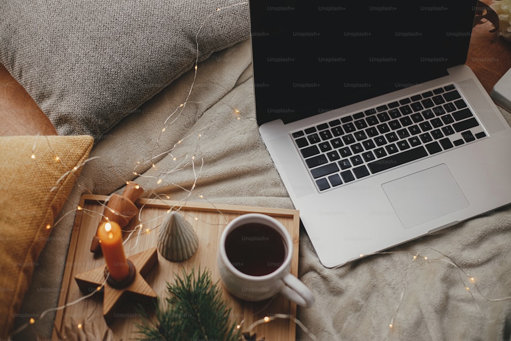 Laptop on soft bed with cup of tea, christmas lights, stars, pine, candle and pillows in scandinavian room. Freelance and blog. Remote work and online. Cozy winter holidays at home
