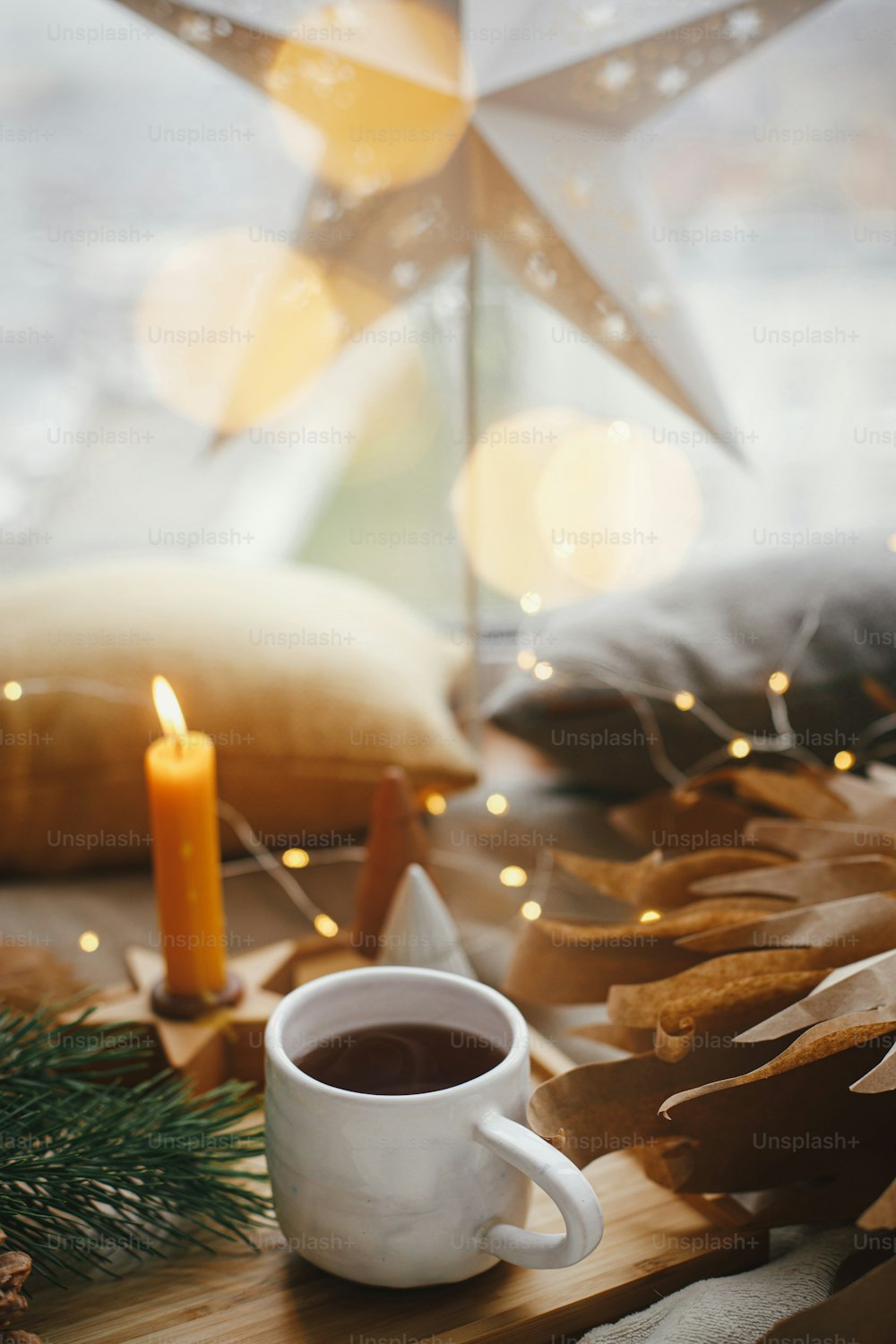 Warm cup of tea on background of illuminated christmas star, christmas lights, pine trees, candle, pillows on bed at window. Cozy scandinavian home. Winter hygge. Atmospheric magic moment