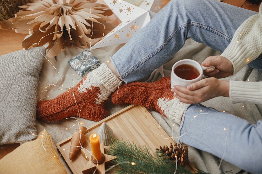 Woman hands with warm cup of tea relaxing on soft bed with christmas stars, golden lights, trees, candle and pillows in scandinavian room.  Cozy moments at home. Winter holidays
