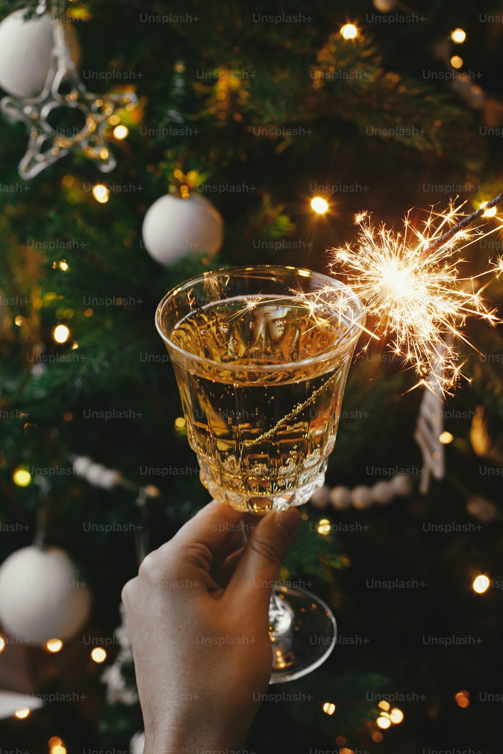Hand haltendes Champagnerglas mit brennender Wunderkerze auf dem Hintergrund der Weihnachtsbaumbeleuchtung. Frohes neues Jahr!! Feuerwerk bengalisches Licht und Getränk in Frauenhand. Atmosphärischer Moment. Silvesterparty