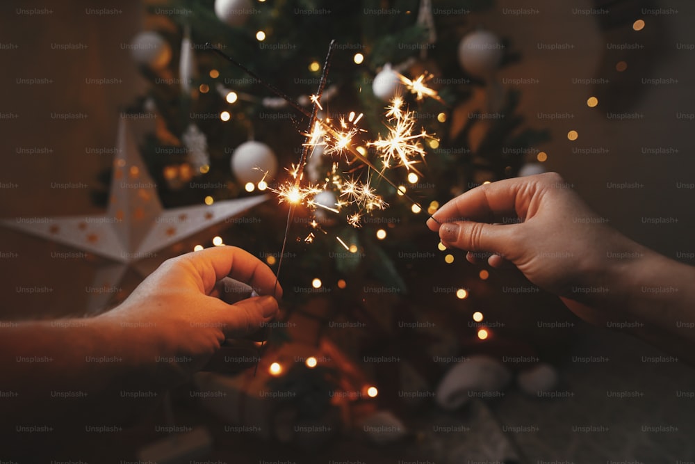 Happy New Year! Hands holding burning sparklers on background of christmas tree in festive scandinavian room. Couple celebrating with fireworks bengal lights. Atmospheric moment. Happy Holidays!