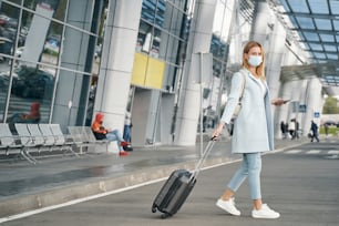 Touriste à la mode dans un masque médical jetable roulant sa valise à roulettes à travers la rue