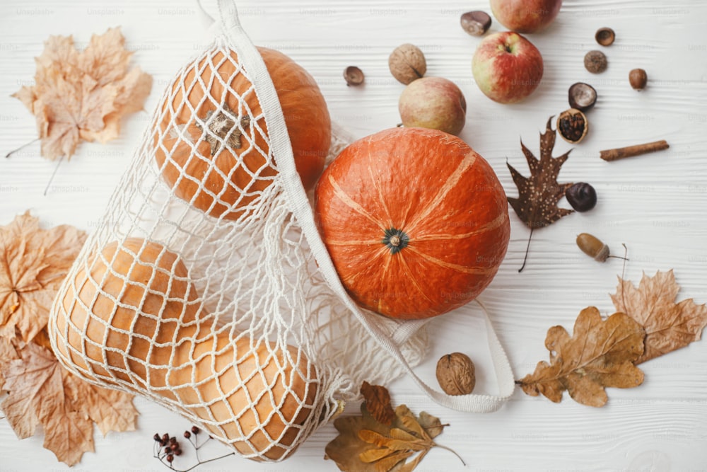 Pumpkins in string bag, apples, nuts and autumn leaves on white wooden background. Autumnal Flat lay. Zero waste concept, plastic free shopping. Harvest. Happy Thanksgiving