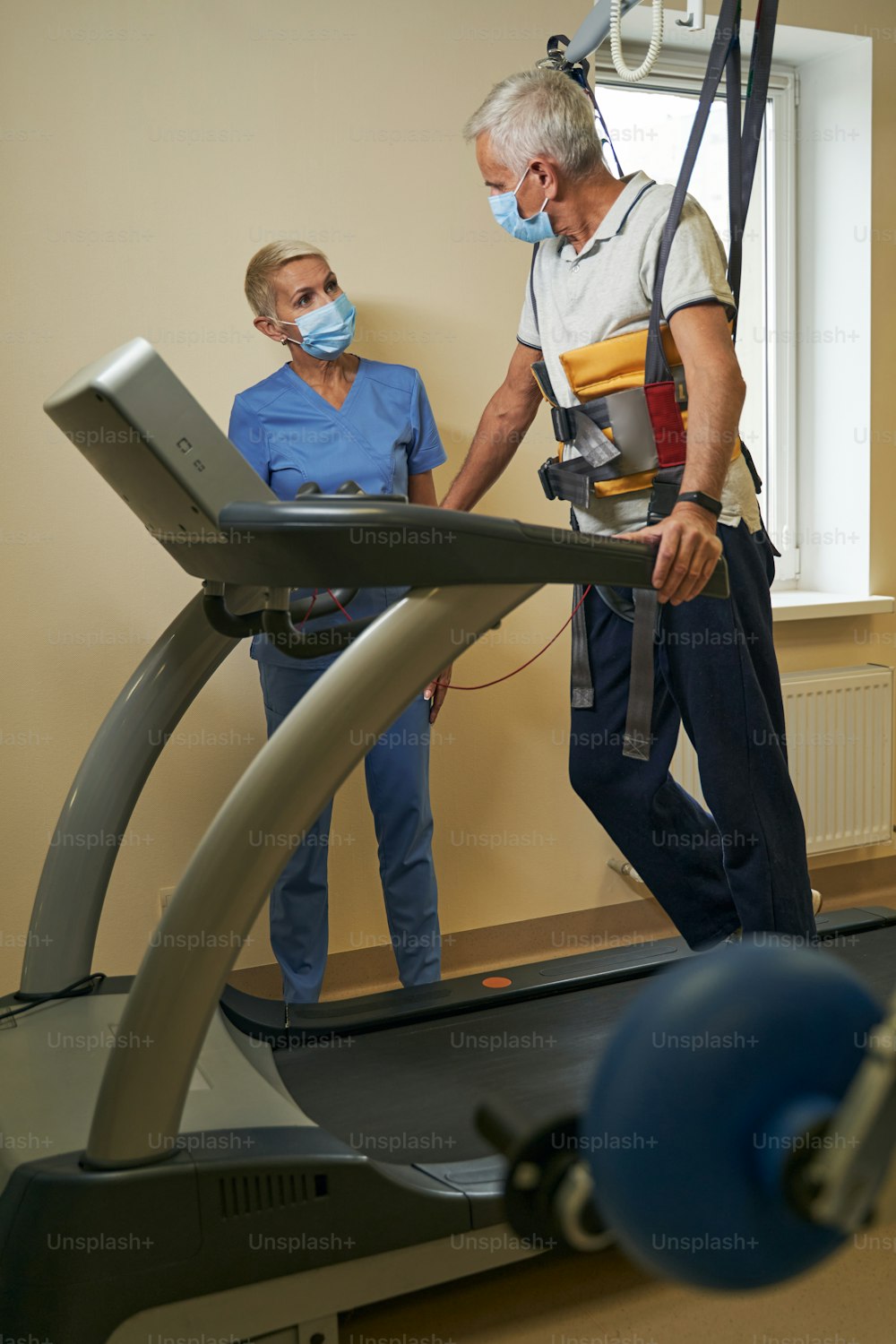 Mature patient on treadmill in face mask using walking harness system for mobilization and movement training in rehab center