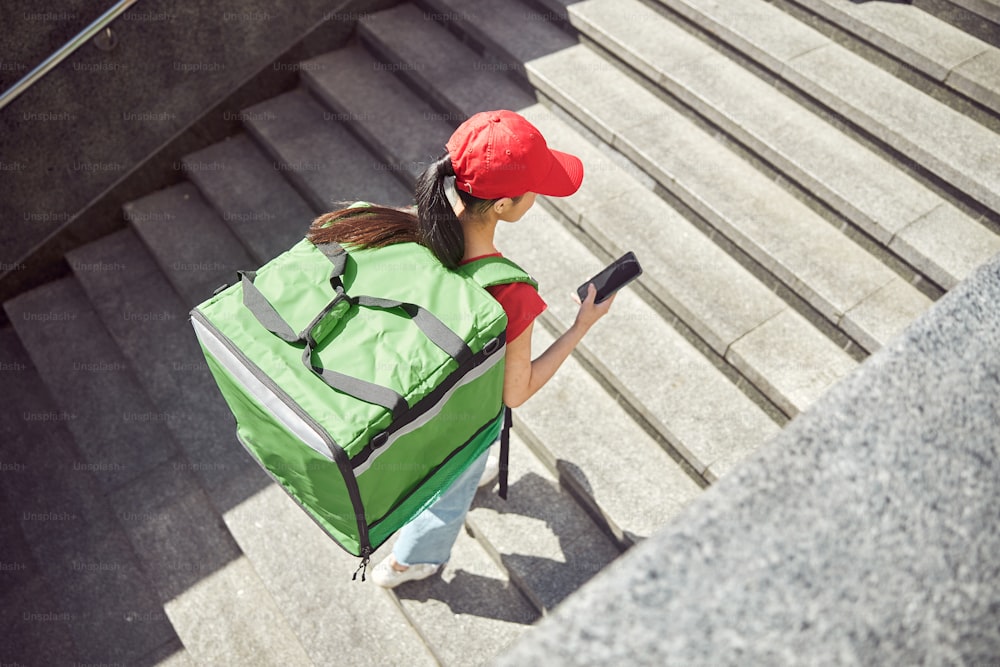 Vista de ángulo alto de una joven repartidora con mochila de comida que navega por la aplicación de mapa en el móvil para encontrar la dirección de entrega al aire libre