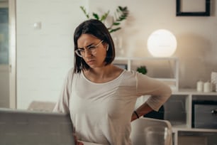 Beautiful woman having back and neck pain while using a laptop and managing her business and working remotely from home