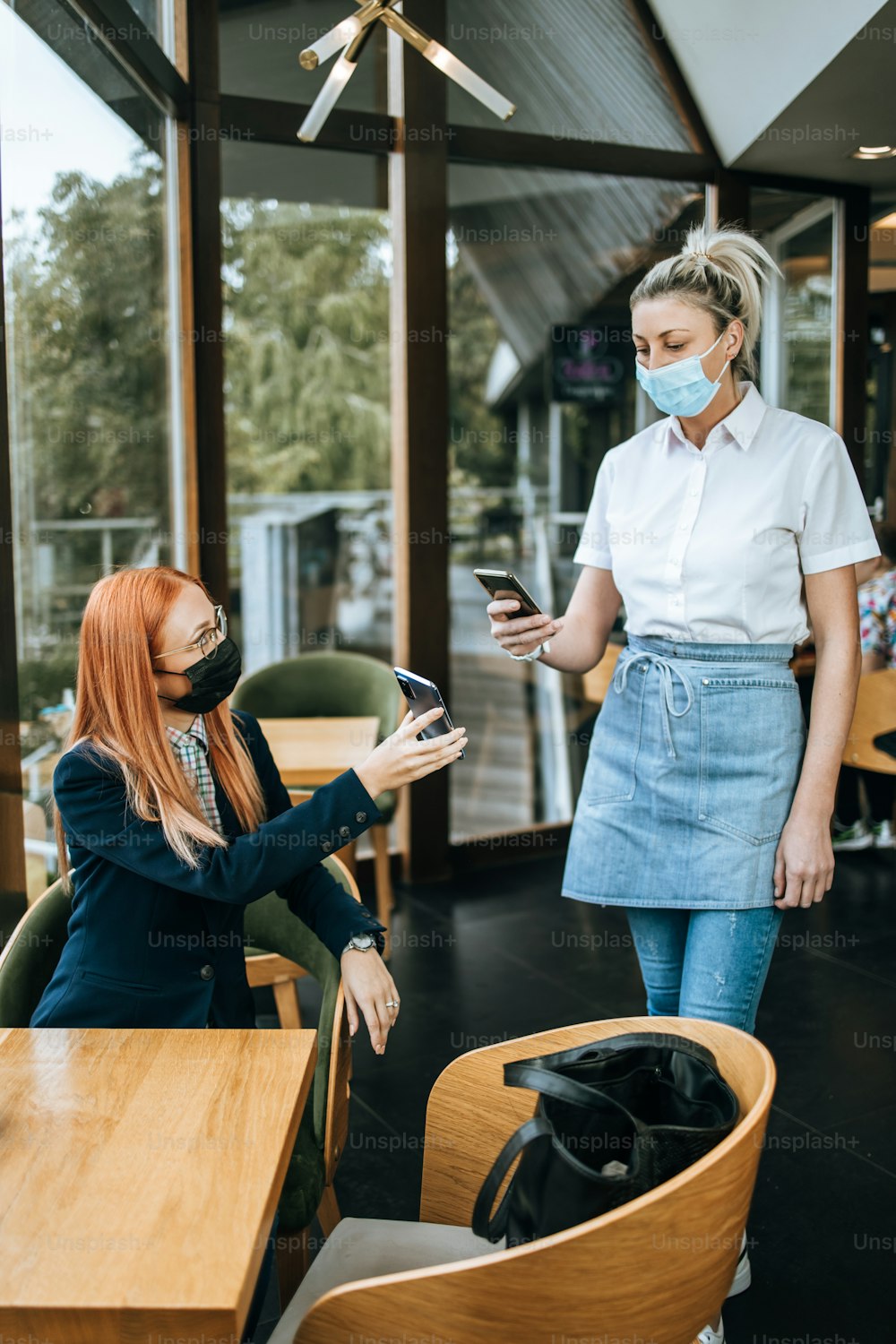 Young attractive business woman sitting in a restaurant or cafe bar and showing digital Covid-19 immunization pass certificate to the security guard. Pandemic and global security measures concept.