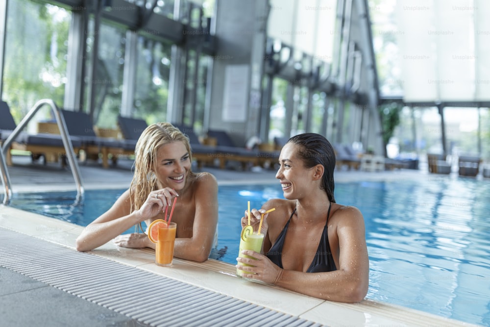 Amigos junto a la piscina relajándose tomando bebidas saludables. Mujeres jóvenes sensuales que se relajan en la piscina del balneario, piscina cubierta del balneario