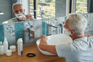 Alter Mann starrt im Badezimmerspiegel auf sein Gesicht und rasiert Gesichtsbehaarung mit Rasierklinge