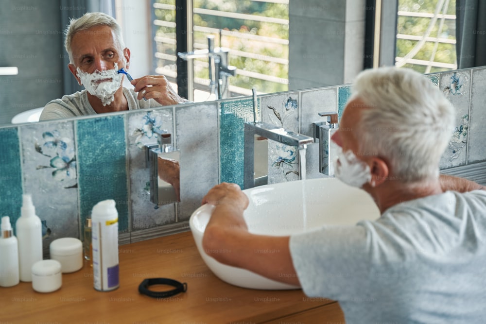 Homem idoso olhando para seu rosto no espelho do banheiro e raspando pelos faciais com lâmina de barbear