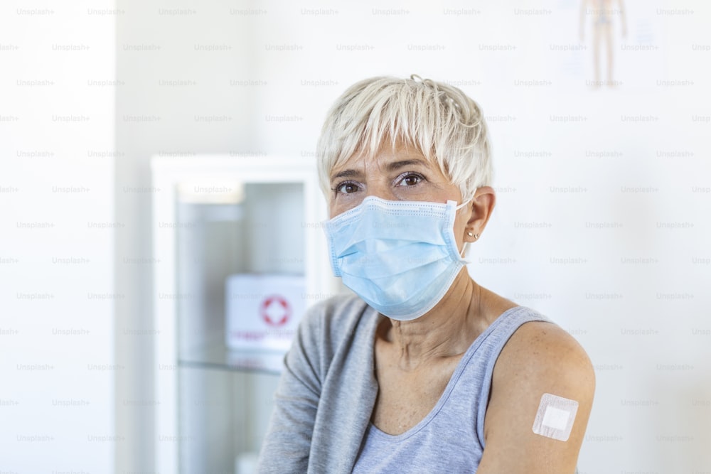 ワクチン接種後に包帯を巻いた腕を示す防護マスクを着けた高齢女性のポートレート。コロナウイルスのワクチン接種を受けた後、座っている成熟した女性。