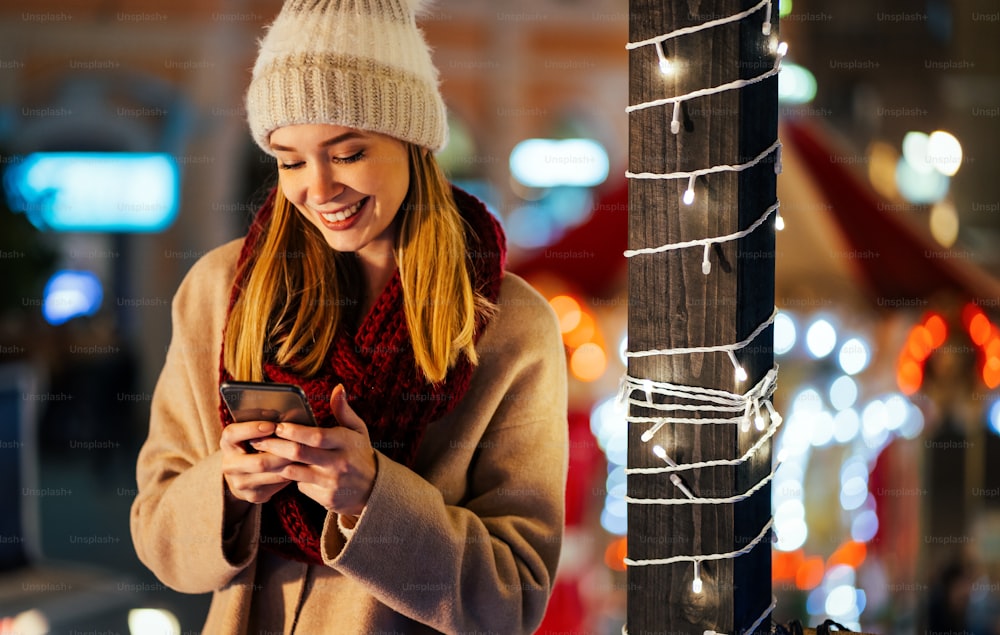 Retrato da mulher jovem bonita que usa seu telefone móvel na rua com a decoração do Natal no fundo