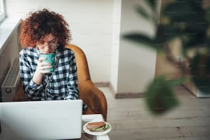 Mulher encaracolada está bebendo um café com hambúrguer enquanto trabalha em um laptop de casa