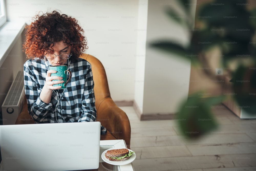 Curly woman is drinking a coffee with burger while working at a laptop from home
