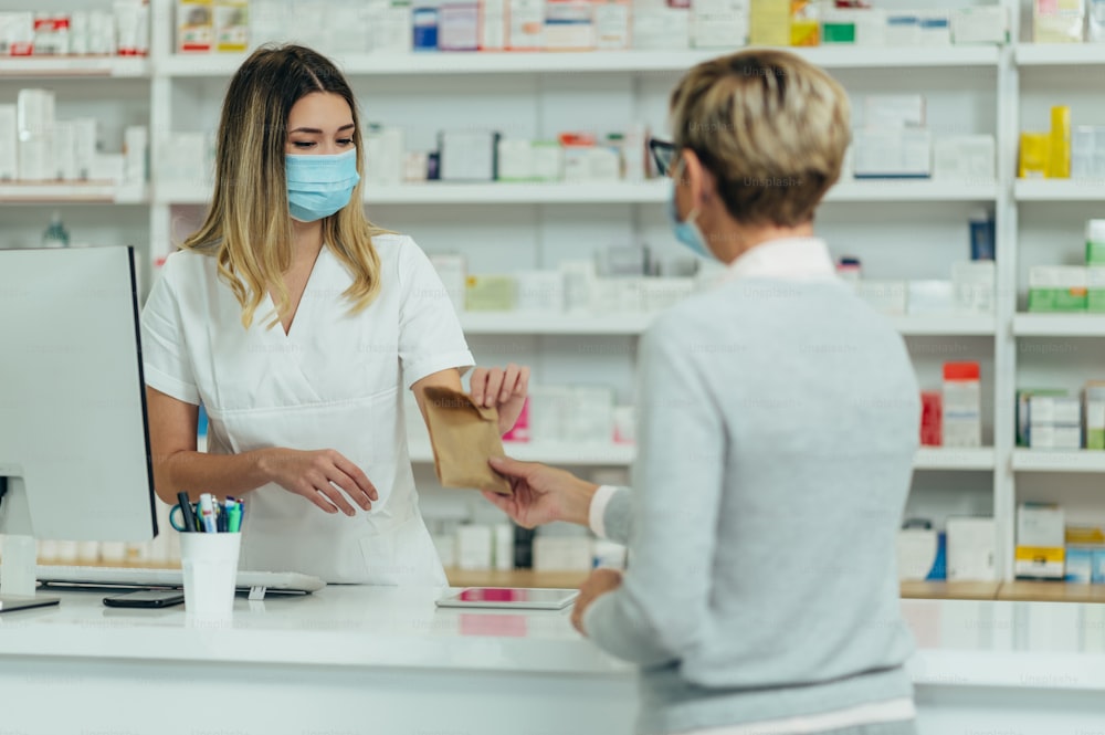 Farmacista donna che indossa una maschera protettiva e serve un paziente cliente in una farmacia e confeziona farmaci in un sacchetto di carta