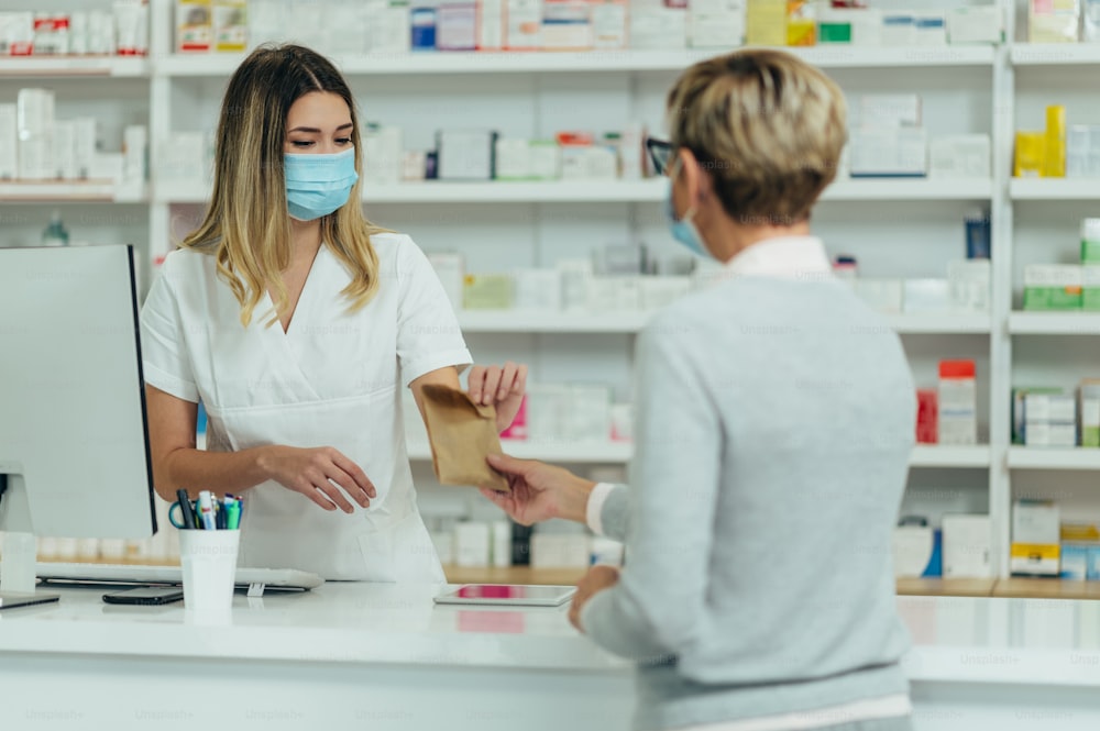 Female pharmacist wearing protective mask and serving a customer patient in a pharmacy and packing drugs in a paper bag
