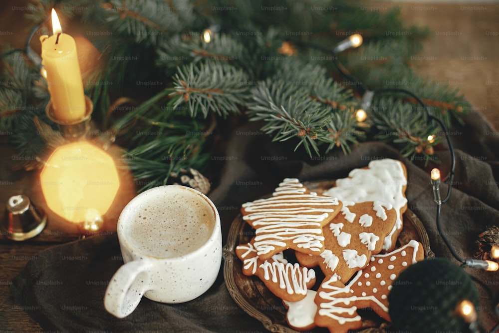 Weihnachtslebkuchenplätzchen, Kaffee in stilvoller weißer Tasse, Tannenzweige, warme Lichter auf Serviette und rustikaler Holztisch. Gemütliches stimmungsvolles Bild. Winterzeit. Frohe Feiertage!