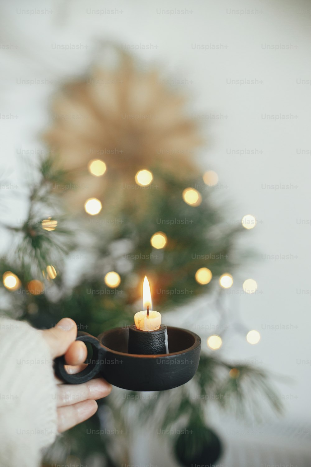 Atmospheric hygge winter home. Hand in cozy sweater holding vintage candlestick with burning candle on background of warm lights, fir branches, sweden star in festive scandinavian room. Xmas advent