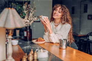 Smiling young woman with curly hair wearing vintage style enjoy tea at the cafe. Selective focus on the woman.