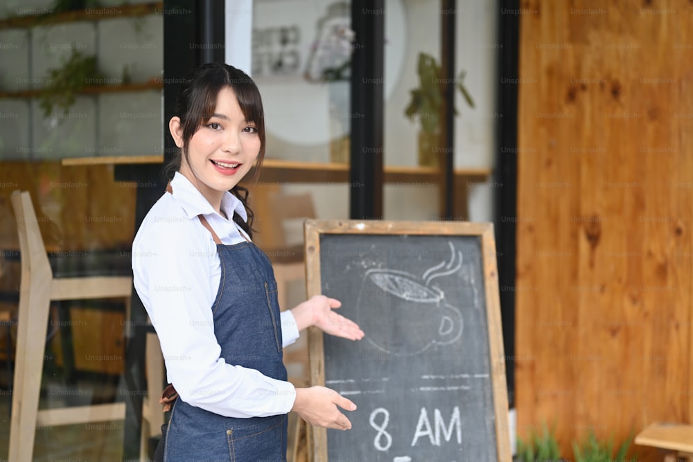 Mulher sorridente dona de cafeteria de pé na porta com um quadro negro.