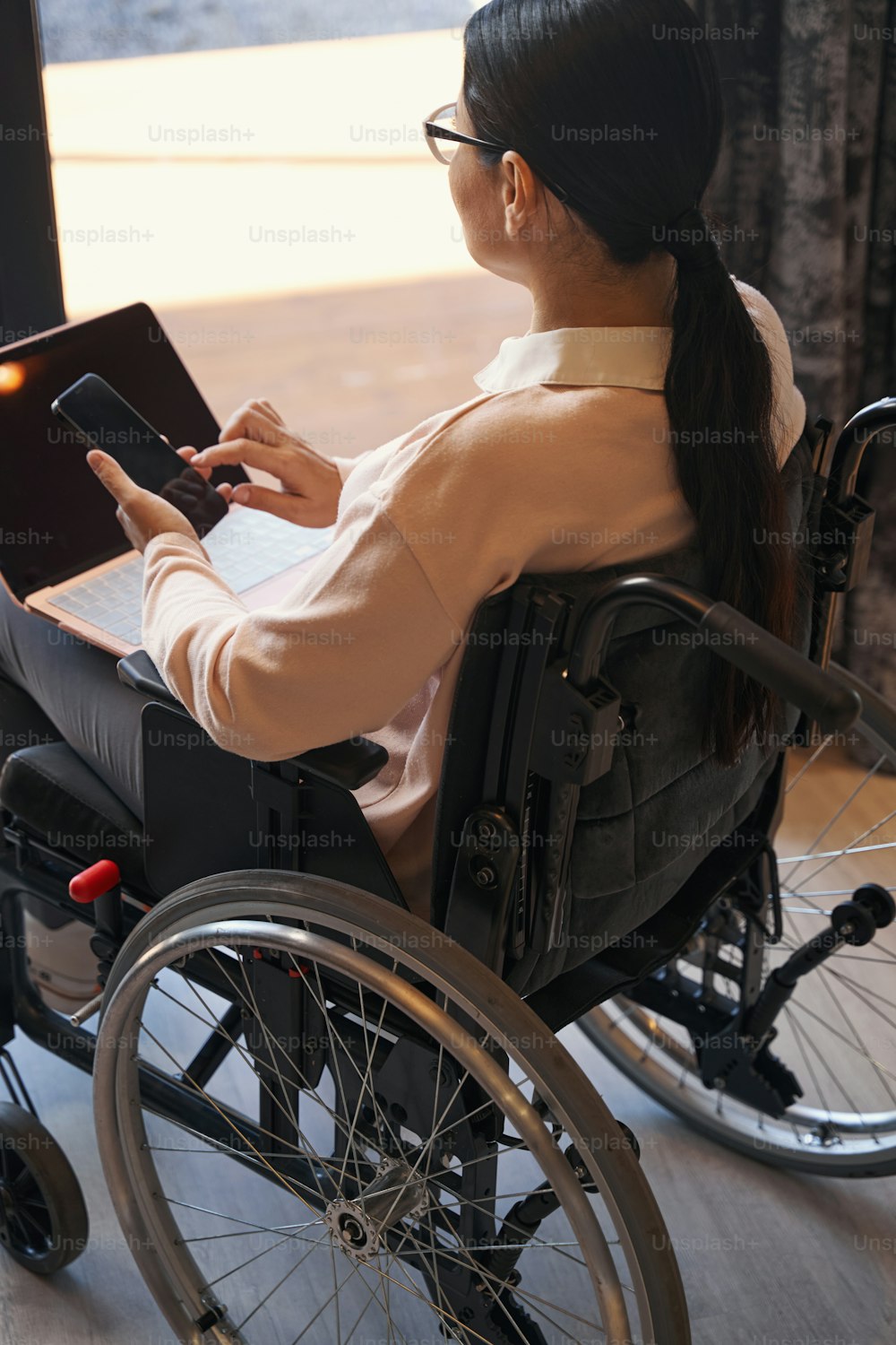 Top view of female sitting in wheelchair and looking out of window while messaging on smartphone and working on laptop