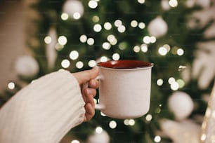 Hand in Pullover hält stilvolle Tasse auf dem Hintergrund des Weihnachtsbaums in Lichtern im Abendzimmer. Platz für Text. Keramiktasse mit Tee in weiblicher Hand im festlichen stimmungsvollen Raum. Gemütliche Winterzeit