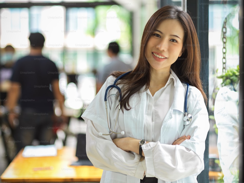Une superbe jeune praticienne, médecin ou médecin asiatique en blouse blanche et stéthoscope croisa les bras près de la vitre.