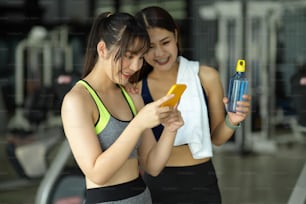 Two of sporty girl standing in fitness studio looking on smartphone screen together after finished workout.