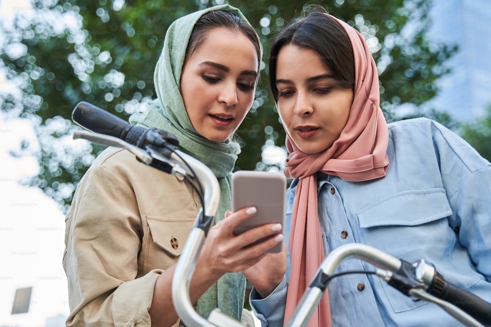 Attractive girls of islamic religion using app near city bicycle station, social network. Low angle view of young millennial women using cellphone. Bike sharing service concept
