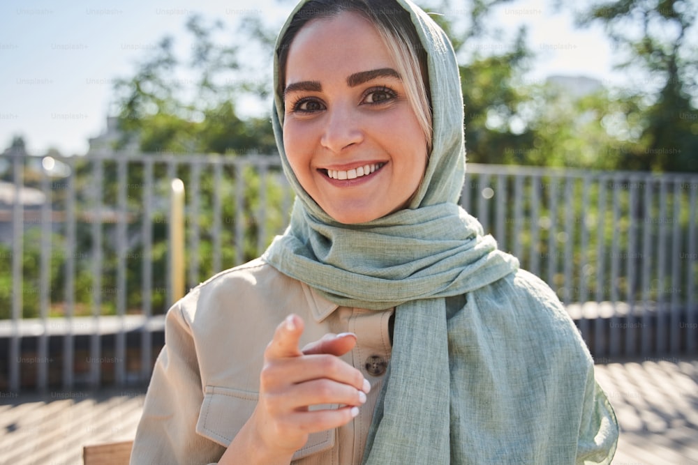 You. Happy modern girl wearing headscarf pointing to the camera with her finger and smiling toothy while chatting with her followers. Stock photo