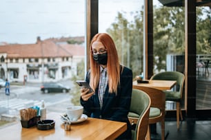 Young elegant business woman sitting in a restaurant or cafe bar and using her smart phone. She is wearing protective face mask against virus disease. Coronavirus and new normal concept.