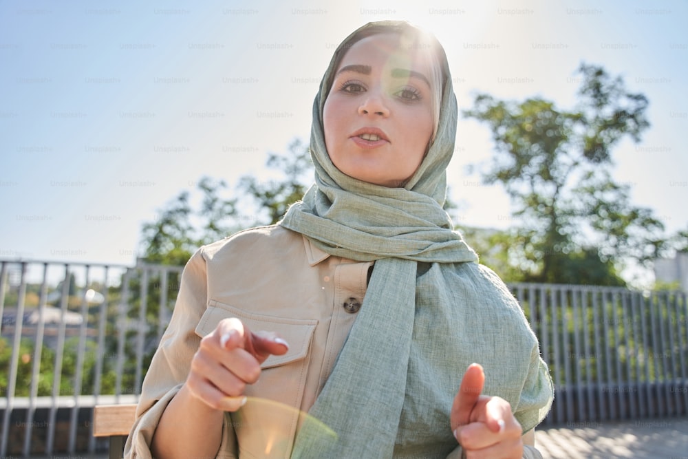 Vue en contre-plongée de la femme arabe confiante pointant avec ses doigts vers la caméra tout en diffusant pour ses abonnés dans la rue