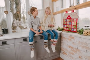 家の形をしたクリスマスの手作りアドベントカレンダーの近くのキッチンに座って楽しんでいる幸せな男の子と女の子。